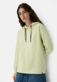 Jasnozielona bluza z kapturem z miękkiego materiału, wyposażona w sznurki i ściągacze przy mankietach. W połączeniu z dżinsową spódnicą, o minimalistycznym wzornictwie.