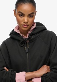 Felpa nera con cappuccio, dotata di coulisse, chiusura con zip e logo ricamato. Il colletto rosa si intravede sotto lo strato esterno nero.