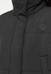 Μαύρο επενδεδυμένο μπουφάν με ψηλό γιακά, που διαθέτει κυκλικό μαύρο λογότυπο στο στήθος και τσέπη στο μπροστινό μέρος. Λείος ματ υφή.