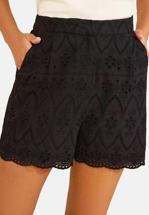 Čierne krátke nohavice s vysokým pásom s kvetinovou dierkovanou výšivkou, prúžkovaným lemom a vreckami, nosené s rukami vreckách.