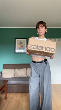 Smeđi kartonski okvir s isprintanim natpisom "zalando" u tamnosivoj boji. Model nosi bijeli kratki top i široke sive hlače, stoji unutra. Iza njega je zelena zid.