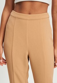 Beige op maat gemaakte broeken met een gladde textuur, hoge taille en verticale naden. De stof lijkt lichtgewicht en iets rekbaar.