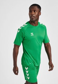 Groen sportshirt en -shorts van glad materiaal. Beschikt over witte chevrons op de mouwen en onderrand, met het 'hummel' logo op de borst.