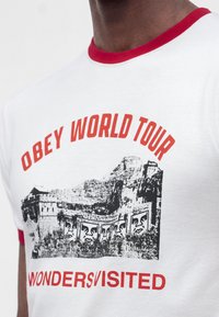 Άτομο που φοράει λευκό T-shirt με κόκκινο γιακά και μανίκια, τυπωμένο με "OBEY WORLD TOUR" και ένα γραφικό με ένα μνημείο με στυλιζαρισμένα πρόσωπα.