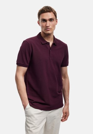 Man met een donkerpaarse korte mouwen polo en lichte broek, staand met één hand in zijn zak tegen een effen achtergrond.