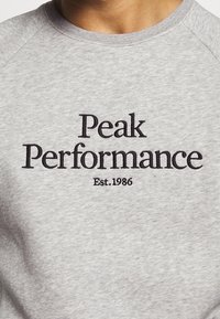 Felpa grigia con il testo ricamato "Peak Performance" in grigio scuro, con "Est. 1986" sotto. Il tessuto ha una texture liscia.