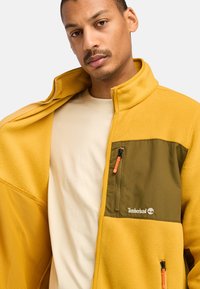 Fleece-Jacke in senfgelb mit einer grünen Nylon-Tasche, durchgehendem Reißverschluss und orangefarbenen Reißverschluss-Zügen. Verfügt über einen hohen Kragen und strukturierten Stoff.