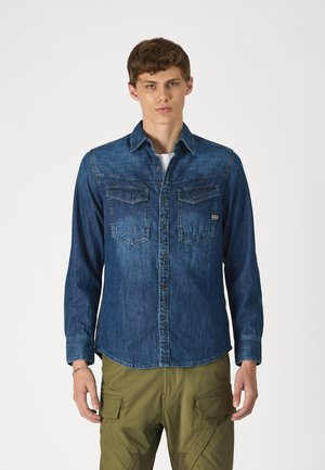 Long-sleeved denim shirt in dark blue, featuring button-up front, two chest pockets, and a classic collar. Smooth, textured fabric.