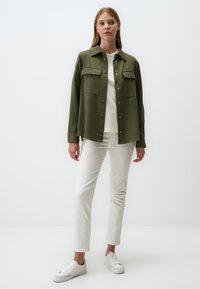 Vrouw die staat en een groene button-up jas over een wit shirt, witte broek en witte sneakers draagt, tegen een effen achtergrond.