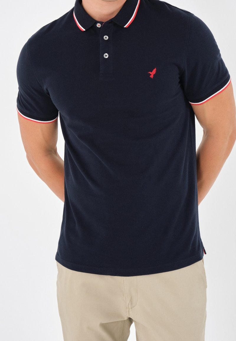 Polo bleu marine en coton, avec un logo d'oiseau rouge, un col à rayures rouges contrastantes et des manches courtes avec des détails accentués.
