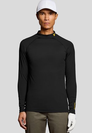 Man draagt een zwart, lang mouws sportshirt met een geel logo op de kraag en een witte golfhandschoen, gecombineerd met een bruine broek en een witte pet.