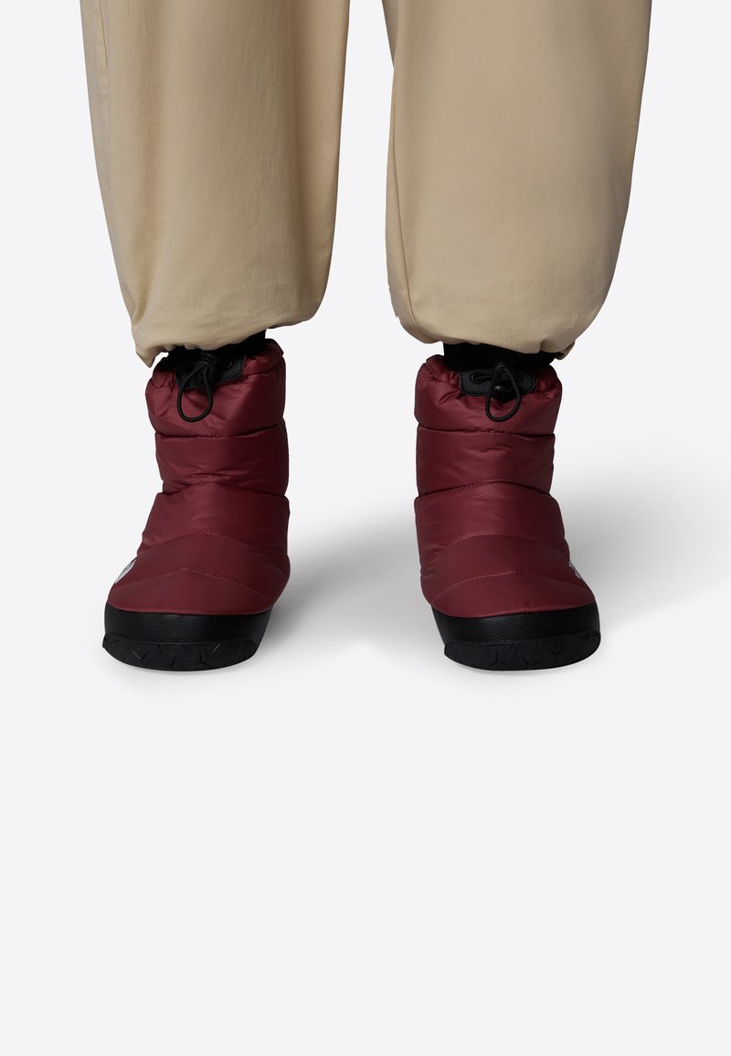 Bottes isolées rouges avec un design enflé, tissu texturé et semelles en caoutchouc noir. Dotées d'une fermeture élastique pour un ajustement sécurisé. Pantalon beige clair visible.