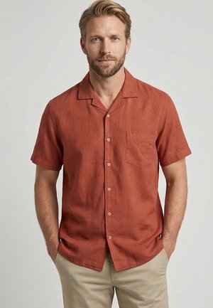 Blondeman met baard die een kort mouwen roestkleurig overhemd met knopen en beige broek draagt, staand met handen in de zakken.