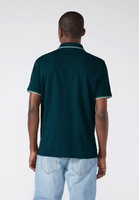 Polo shirt verde scuro con un colletto a contrasto bianco e verde, maniche corte e una texture liscia. Indossato con jeans blu chiaro.