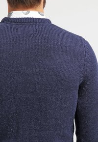 Marinblå stickad tröja med en strukturerad yta, med rund halsringning och ribbade ärmslut. Bakifrån avslöjas en slät, kontinuerlig design.