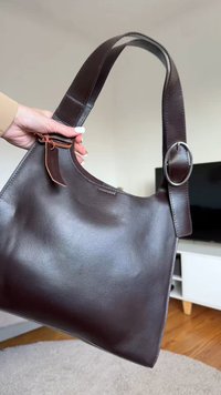 Brun läderhandväska med bred axelrem, rundad form, en dekorativ tagg och en rundad metallringsdetalj. Mjuk textur, minimalistisk design.