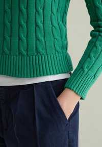 Maglione verde lavorato a maglia con motivi a treccia, polsini a coste e fondo dritto, abbinato a pantaloni blu scuro.
