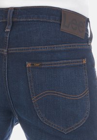 Jeans de denim azul oscuro con costuras naranjas, que presentan una etiqueta de cuero en la cinturilla y un solo bolsillo posterior con una pequeña etiqueta de la marca.