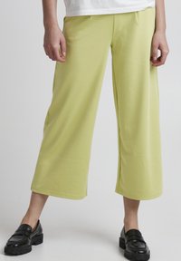 Heledad kollastest laiad püksid, mis on valmistatud pehmest kangast ja millel on sile tekstuur, keskmise kõrgusega talje ja peened volditud detailid. Kanda mustade loafersitega.