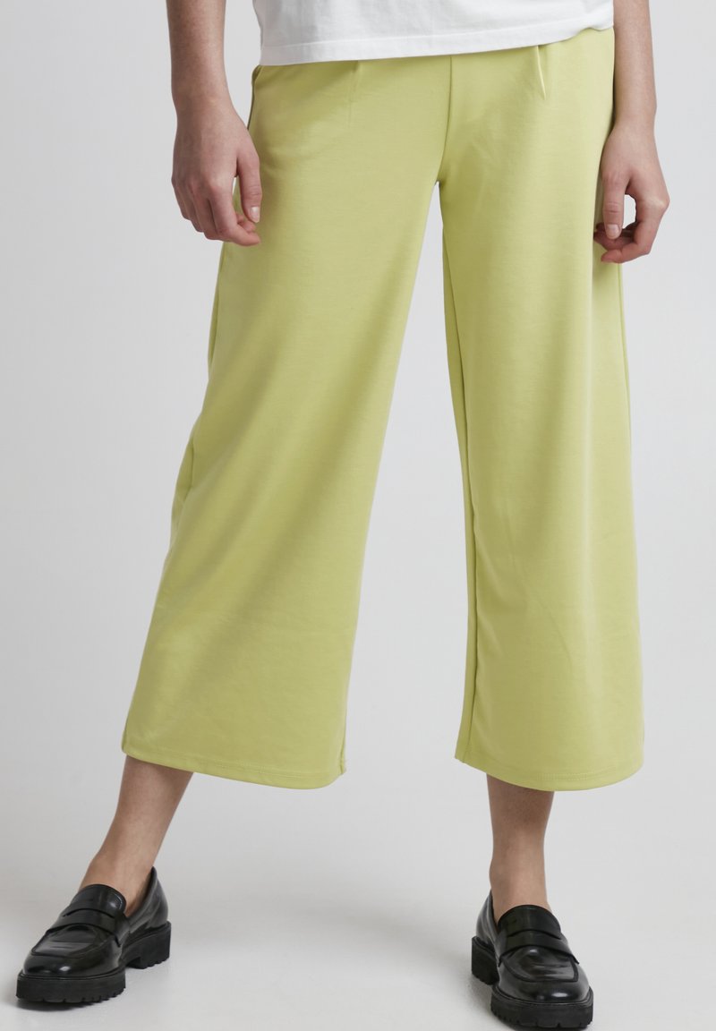 Heledad kollastest laiad püksid, mis on valmistatud pehmest kangast ja millel on sile tekstuur, keskmise kõrgusega talje ja peened volditud detailid. Kanda mustade loafersitega.