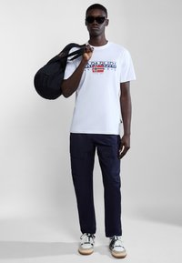 Tricou alb cu un grafic albastru și roșu, pantaloni scurți navy, rucsac negru și adidași albi cu accente maronii. Bărbat purtând ochelari de soare.