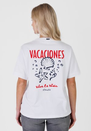 Femme blonde portant un t-shirt blanc avec le texte rouge « VACACIONES » et « Viva La Vida » ainsi que des illustrations noires sur le thème de la mer au dos.