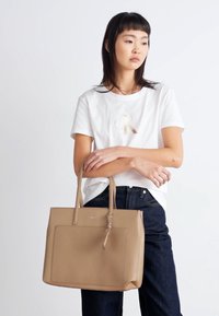 Tan synthetic tote bag with vertical stitching, flat shape, and dual handles. Model wearing a white T-shirt and dark jeans holds the bag.