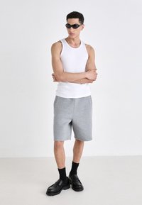 Bílý bezrukávový top, šedé šortky, černé robustní mokasíny a černé sluneční brýle. Model pózuje se zkříženýma rukama na jednobarevném pozadí.