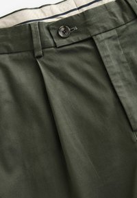 Pantalon vert olive avec une taille boutonnée, passants de ceinture et une doublure intérieure écrue partiellement visible avec une bande bleu marine.