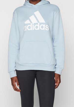 Personne portant un sweat à capuche Adidas bleu clair avec poche avant et des leggings noirs, debout devant un fond uni clair.