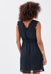 Robe noire avec une texture à petits pois, décolleté en V, bordures en dentelle aux épaules, taille cintrée et jupe évasée.