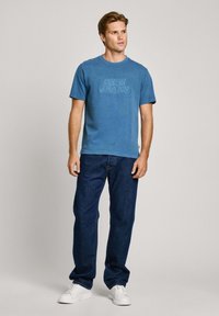 Mėlynas medvilninis marškinėlis su iškiliais "PEPE JEANS" užrašais, derinamas su tamsiais džinsais ir baltais sportbačiais. Kasdieniam derinimui ir stiliui.