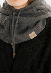 Vrouw met een dikke grijze gebreide sjaal met houten kralen over een zwart textuurjack, met een "Knit Factory" label zichtbaar.