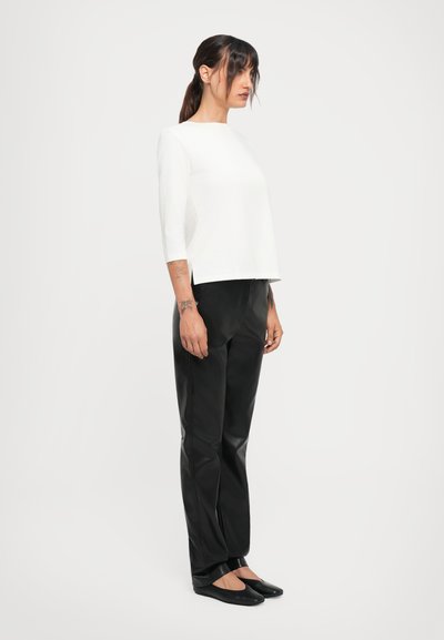 Woman standing sideways wearing a white long-sleeve top, black leather pants, and black flats on a plain white background.
