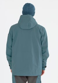 Vedeněodolná teal bunda s kapucí, elastickými manžetami a rovnou siluetou. Nositelná přes šedé kalhoty, které vykazují hladkou texturu a minimální švy.