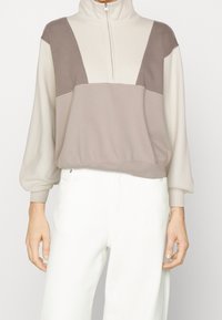 Suéter ligero de bloques de color con un alto collar y cierre de media cremallera. Presenta materiales suaves en tonos beige y topo, y mangas sueltas.
