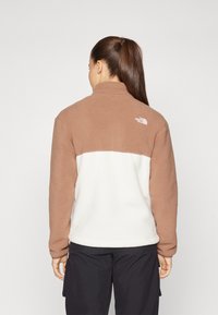 Fleece jakna v rjavi in beli barvi, z visokim ovratnikom in logotipom na hrbtu. Ima mehak material in sproščen kroj.