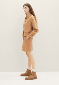 Veste en velours côtelé marron avec poche poitrine, assortie à une jupe. La mannequin porte des bottes marron à lacets et des chaussettes beiges. Fond neutre.