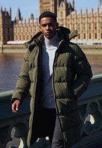 Olivově zelený péřový kabát se strukturovanou kapucí, pod ním šedý žebrovaný rolák a tmavé kalhoty; má polstrovanou texturu a zipové kapsy.