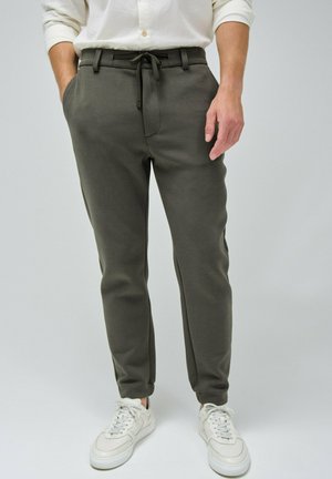 Pantalones jogger verde oliva hechos de tela suave con un diseño tapered, cinturilla ajustable con cordón y bolsillos laterales, combinados con zapatillas blancas.