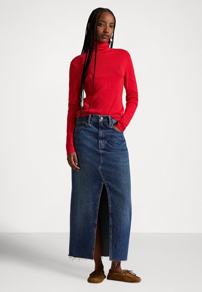 Žena s dlhými vrkočmi, oblečená v červenom roláku, členkovej džínsovej sukni s predným rozparkom a hnedej mokasínovej obuvi.