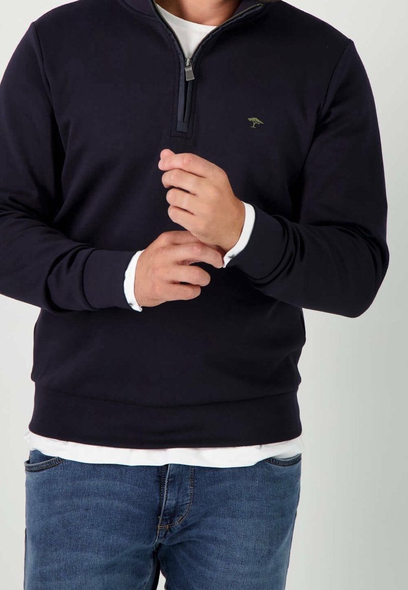 Homme ajustant la manche d'un pull noir à fermeture éclair au quart, par-dessus une chemise blanche, portant un jean bleu, sur un fond clair uni.