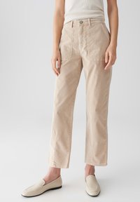 Eine Person trägt beiges, gerade geschnittenes Cord-Hosen, ein weißes Tanktop und cremefarbene Loafers und steht vor einem schlichten, hellen Hintergrund.