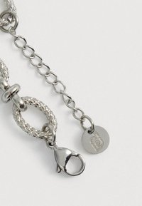 Tuvu redzama sudraba ķēdes kaklarota ar aizdari, kurai ir teksturēti posmi un mazs apaļš birka ar gravētu logotipu uz baltas fona.