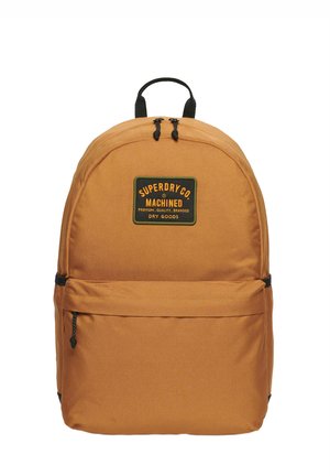 Mochila de tela naranja, de forma redondeada, con un bolsillo frontal con cremallera. Presenta un parche de logo negro y tiradores de cremallera negros. Los detalles incluyen un asa en la parte superior.