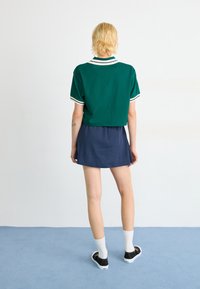 Polo vert court avec col et poignets blancs, associé à une mini-jupe marine. Le modèle porte des chaussures noires et des chaussettes blanches. Le fond est uni.