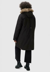 Black parka coat with a fur-trimmed hood, drawstring waist, and snap-button cuffs, paired with black cropped pants and chunky black boots.