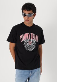 Tricou negru din bumbac cu un mare logo roșu și alb "TOMMY JEANS", un grafic cu un tigru și detalii cu lauri. Modelul poartă ochelari de soare gri.