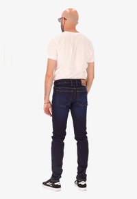 Homme à tête rasée portant des lunettes de soleil noires, un t-shirt blanc, un jean bleu marine et des baskets noires, se tenant de dos sur fond blanc.