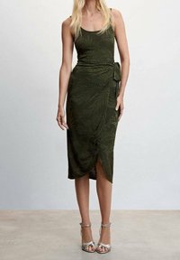 Robe portefeuille sans manches vert olive avec un motif de feuilles, détail noué sur le côté, coupe au genou et fente à l'avant. Des escarpins argentés à lanières sont portés.